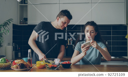 Attractive couple in the kitchen at home. Curly girl playing video game on smartphone while her boylfriend cooking breakfast Attractive couple in the kitchen at home. Curly girl playing video game on smartphone while her boylfriend cooking breakfast 92725109