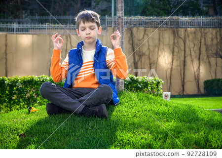 Caucasian boy sitting on fresh green grass field and make meditation dressed in casual clothes, happy kid enjoying peaceful summer weekend outdoors. Relaxation inspiration concept Caucasian boy sitting on fresh green grass field and make meditation dressed in casual clothes, happy kid enjoying peaceful summer weekend outdoors. Relaxation inspiration concept 92728109