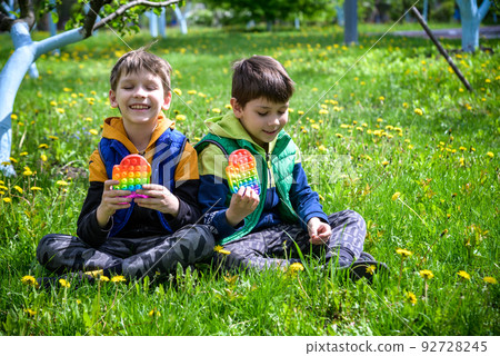 children playing with colorful toys Pop It. Boys on vacation. Tired of games, relaxing on the grass. Summer time for children, two boys friends at reboot together children playing with colorful toys Pop It. Boys on vacation. Tired of games, relaxing on the grass. Summer time for children, two boys friends at reboot together 92728245