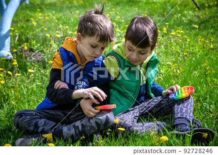 children playing with colorful toys Pop It. Boys on vacation. Tired of games, relaxing on the grass. Summer time for children, two boys friends at reboot together 92728246