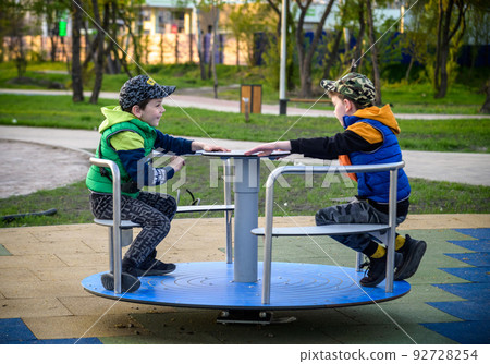 Brightly dressed twin brothers play on a carousel in an autumn park amid fallen yellow leaves. They are two years old Brightly dressed twin brothers play on a carousel in an autumn park amid fallen yellow leaves. They are two years old 92728254