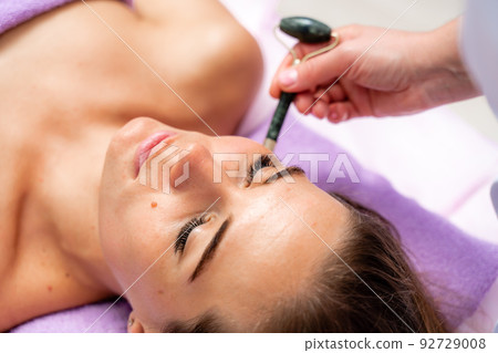 Relaxing massage. Side view european woman getting massage with jade face roller gouache in spa salon Relaxing massage. Side view european woman getting massage with jade face roller gouache in spa salon 92729008