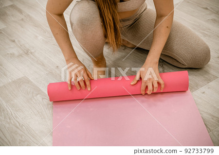 A young woman rolls a pink fitness or yoga mat before or after exercising, exercising at home in the living room or in a yoga studio. Healthy habits, keep fit, weight loss concept. Closeup photo 92733799