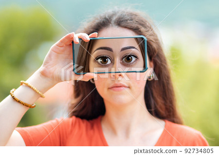 A woman closes her eyes with a smartphone, with a picture of huge cartoon eyes. The concept of hiding identity and fake pages in social networks A woman closes her eyes with a smartphone, with a picture of huge cartoon eyes. The concept of hiding identity and fake pages in social networks 92734805