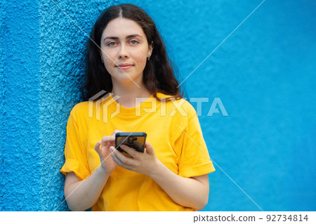 Portrait of a young smiling woman typing on a smartphone. Online surfing concept. Blue wall on the background. Copy space Portrait of a young smiling woman typing on a smartphone. Online surfing concept. Blue wall on the background. Copy space 92734814
