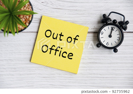 Out of office text on sticky note with alarm clock and plant background. Business concept 92735962