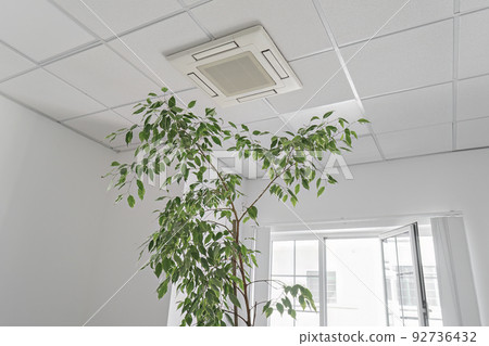 Low angle of assette Air Conditioner on ceiling in modern light office or apartment with green ficus plant leaves. Indoor air quality 92736432