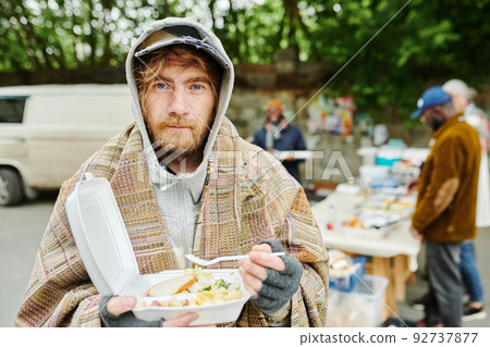 Portrait of bearded homeless man in warm clothing looking at camera while eating food outdoors during charity 92737877