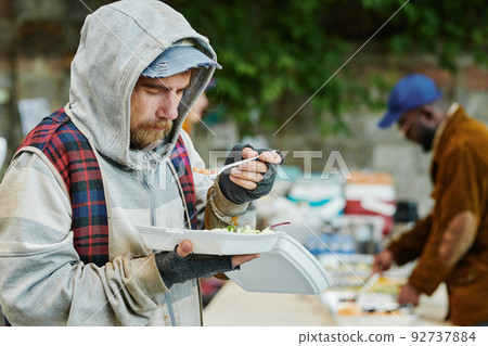 Homeless man in warm torn clothing eating food outdoors giving by volunteers during charity Homeless man in warm torn clothing eating food outdoors giving by volunteers during charity 92737884