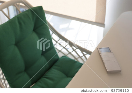 White remote control panel for automatic roller blinds lies on a beige table in the living room. Nearby is a chair with a green pillow. On the window, a roller blind with automatic control.  92739110