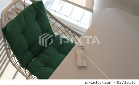 White remote control panel for automatic roller blinds lies on a beige table in the living room. Nearby is a chair with a green pillow. On the window, a roller blind with automatic control.  92739116