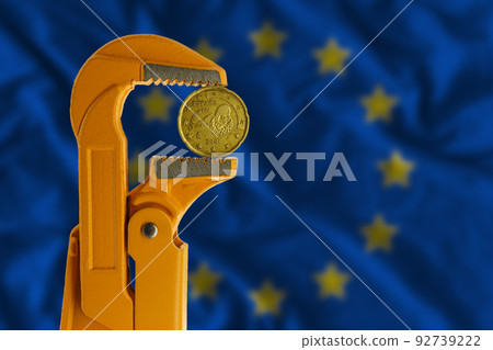 20 euro cent coin held in an orange plumber wrench on the background of the European Flag. Obverse of the coin. 92739222