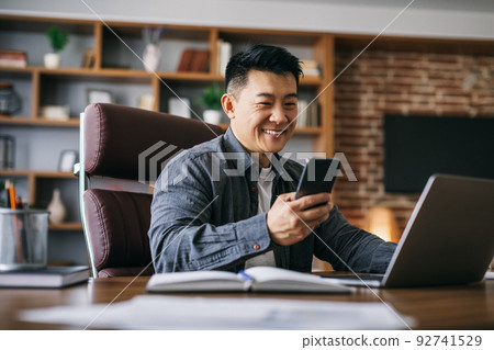 Happy handsome mature asian businessman typing on smartphone at workplace at laptop in home Happy handsome mature asian businessman typing on smartphone at workplace at laptop in home 92741529