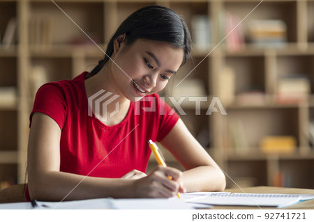 Smiling smart young chinese woman student doing homework, preparing to exam alone in room interior, close up Smiling smart young chinese woman student doing homework, preparing to exam alone in room interior, close up 92741721