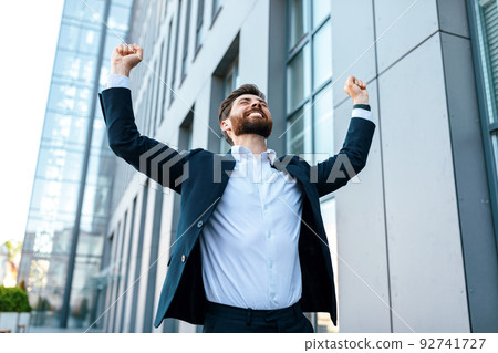 Cheerful attractive young caucasian businessman with beard in suit raises his hands up and rejoices to victory 92741727