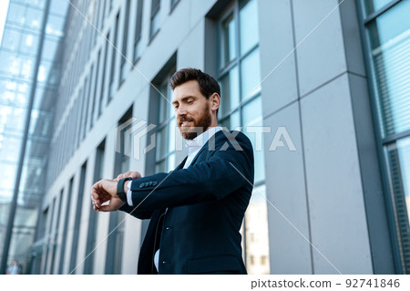 Smiling confident handsome young european male with beard in suit looks at watch, waits client 92741846