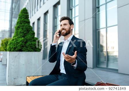 Happy millennial attractive european man with beard in suit calling by phone to client 92741849