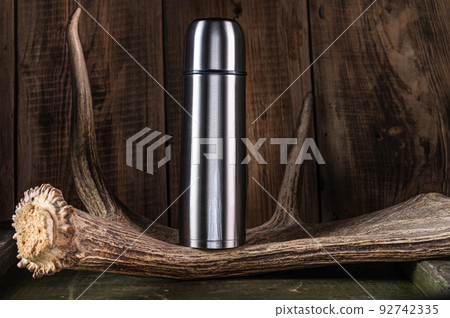 Steel shiny thermos on a wooden background. Thermos on the horn of an elk. Thermos for hunters and tourists. Steel shiny thermos on a wooden background. Thermos on the horn of an elk. Thermos for hunters and tourists. 92742335