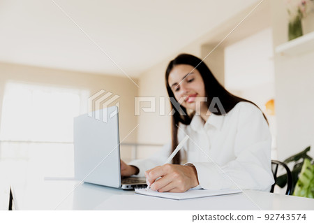 Caucasian business woman sitting at desk, reading from computer, taking notes at notebook. Female student using pc, writing down important information with pencil in note pad. Happy smiling lady Caucasian business woman sitting at desk, reading from computer, taking notes at notebook. Female student using pc, writing down important information with pencil in note pad. Happy smiling lady 92743574