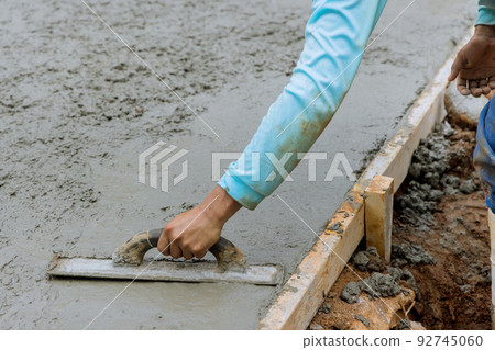 Mason worker is holding steel trowel and smoothing plastering new sidewalk on wet freshly poured concrete 92745060