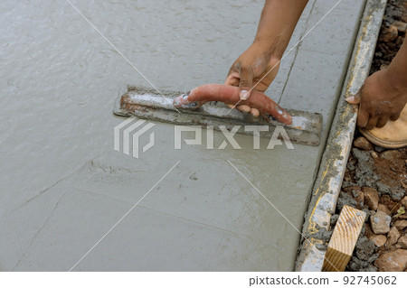 A worker is working on a new sidewalk that has just been poured on wet concrete while holding a steel trowel A worker is working on a new sidewalk that has just been poured on wet concrete while holding a steel trowel 92745062