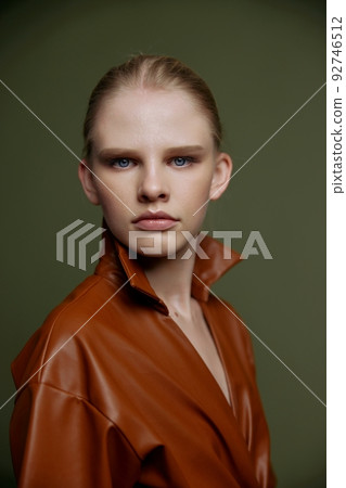 Stylish serious young blonde girl with blue eyes with evening makeup in a leather dress looks at the camera posing isolated. Studio portrait on a green background. Fashion offer 92746512