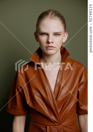 Seasonal Sale offer. Confidence blond young lady in trendy leather trench coat dress looks with blue eyes at camera presenting evening makeup posing isolated on olive green studio wall background 92746514
