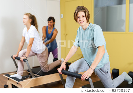 Young boy looking in camera while training pilates in gym. Their trainer hispanic woman helping girl with exercises on pilates reformer in background. Young boy looking in camera while training pilates in gym. Their trainer hispanic woman helping girl with exercises on pilates reformer in background. 92750197