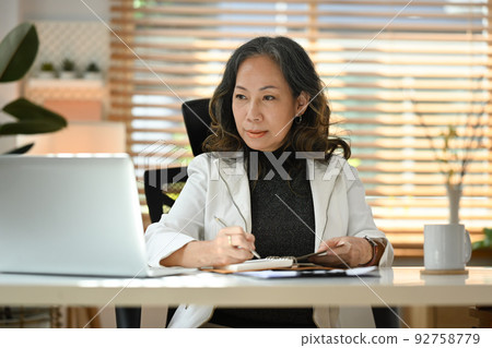 Focused middle aged woman entrepreneur reading online information on laptop and making notes on notebook 92758779