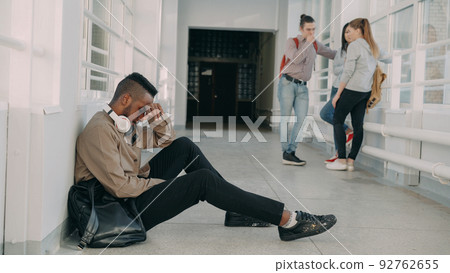 View of upset serious african-american male student sitting on floor in white spacious university corridorwhile his groupmates are standing communicating. View of upset serious african-american male student sitting on floor in white spacious university corridorwhile his groupmates are standing communicating. 92762655
