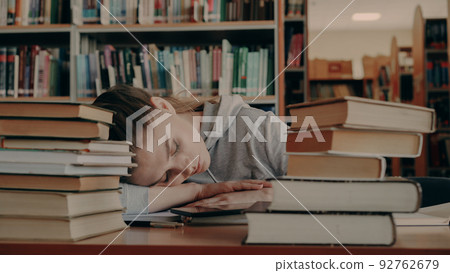 Young beautiful caucasian female student is sleeping with head on table surrounded by piles of books and papers, phone is lieing in front of her, she is in large university library Young beautiful caucasian female student is sleeping with head on table surrounded by piles of books and papers, phone is lieing in front of her, she is in large university library 92762679