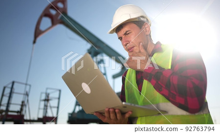 oil industry. engineer next to an oil rig. oil production business finance concept. gas production. a worker with a laptop monitors oil sun production next to an station. a worker in hard hat works 92763794