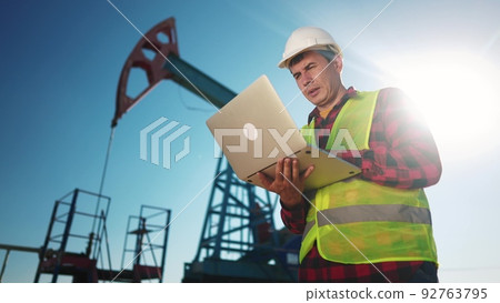 oil industry. engineer next to an oil rig. oil production business finance concept. gas production. a worker with a laptop monitors oil production next to an station. a worker sun in hard hat works 92763795