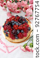 Berry birthday cake. Cheesecake with cherries, raspberries, currants and blueberries. Cake, pink candles and red flowers on a white background. Photo for stories. 92767754