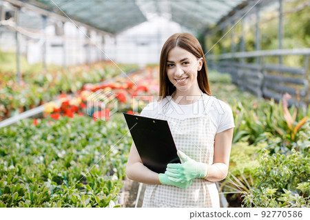Portrait of gardener woman at work in greenhouse with notebook examines the growing flowers in greenhouse. Home gardening, love of plants and care. Small business. 92770586