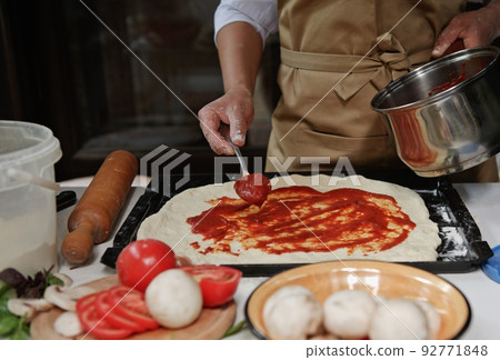 Cropped Italian housewife in beige chef's apron spreading freshly made tomato sauce on rolled out dough in pizza pan 92771848