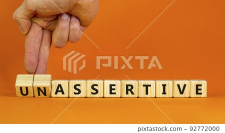 Assertive or unassertive symbol. Businessman turns wooden cubes and changes concept words Unassertive to Assertive. Beautiful orange background. Business assertive or unassertive concept. Copy space. 92772000