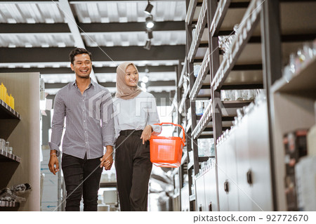 Asian muslim young couple carrying baskets walking between shelves Asian muslim young couple carrying baskets walking between shelves 92772670