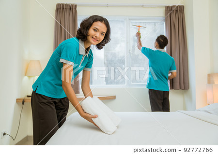 Smiling female housekeeper in turquoise uniform putting towel on bed 92772766