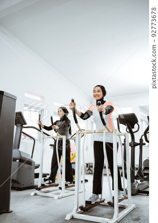 woman with hijab exercising with friend on static elliptical cycle machine woman with hijab exercising with friend on static elliptical cycle machine 92773678
