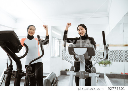 excited muslim woman exercising with friend on static elliptical cycle machine 92773801