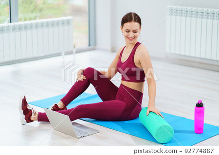 a beautiful sports woman in a burgundy tracksuit with laptop on a mat in a gym. a beautiful sports woman in a burgundy tracksuit with laptop on a mat in a gym. 92774887