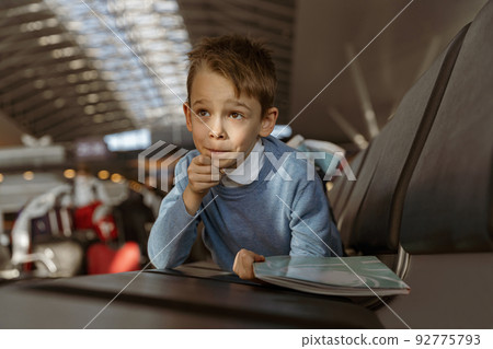 Little boy holding a book in the waiting room at the airport before the flight and looking away Little boy holding a book in the waiting room at the airport before the flight and looking away 92775793