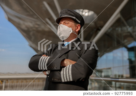 Mature pilot in mask in uniform standing outdoors near airport building Mature pilot in mask in uniform standing outdoors near airport building 92775993