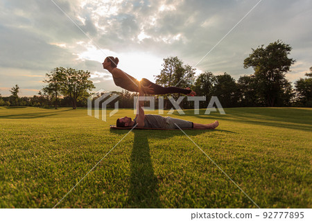 Healthy man lying on grass and balancing woman. Couple doing acrobatic yoga exercise in park Healthy man lying on grass and balancing woman. Couple doing acrobatic yoga exercise in park 92777895