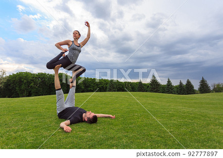 Young couple doing acro yoga in park. Man lying on grass and balancing woman in his feet 92777897