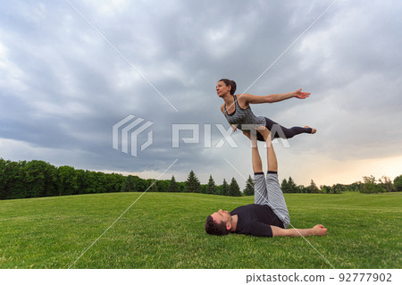 Young couple doing acro yoga in park. Man lying on grass and balancing woman in his feet 92777902