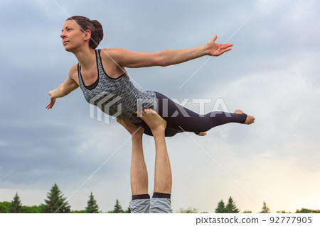 Close up of man lying on grass and balancing woman in his feet. Young couple doing acro yoga in park 92777905