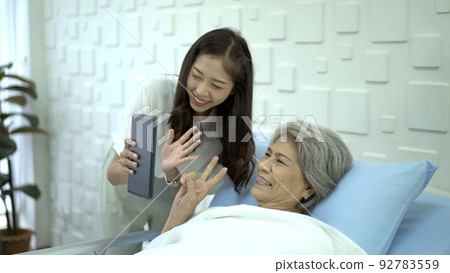 Senior elder Asian female patient resting on the medical bed in hospital and talking to her daughter. daughter visiting  his mother in hospital and let her talk to family on a video call on digital ta 92783559
