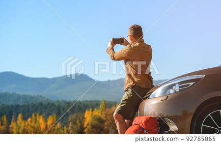 Man sits on car bumper using mobile phone to make picture of beautiful mountains view during summer trekking. Tourist with backpack stop to relax and enjoy nature 92785065